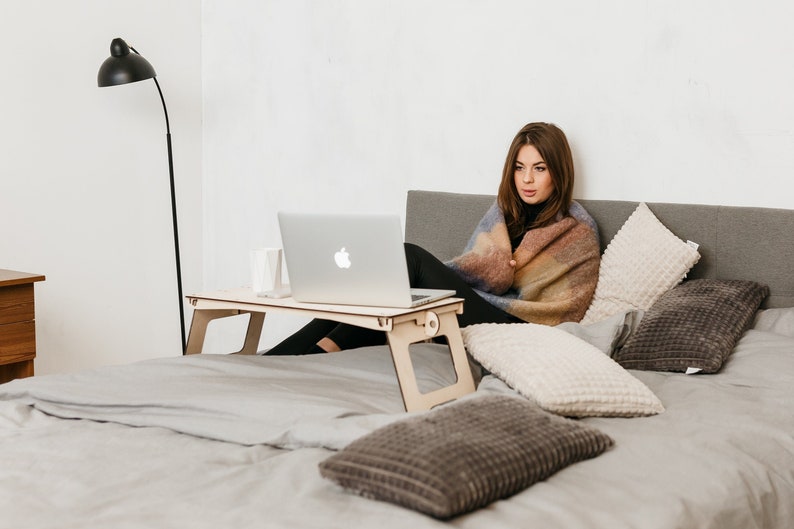 May include: A woman sits in bed with a blanket wrapped around her, using a laptop on a wooden lap desk. The bed has a gray duvet and several pillows.