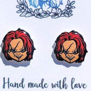 May include: A pair of Chucky-themed earrings on a white card. The earrings feature the character's face with red hair, stitched scars, and a sinister grin. The card also displays the text "Lola's Corner Shop" and "Hand made with love."