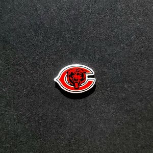 May include: A red and white enamel pin in the shape of the letter "C" with a bear head logo. The bear is red with black details, and the pin has a white border.