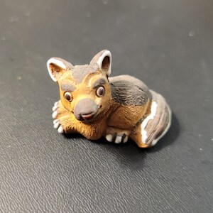 May include: A small, detailed figurine of a resting animal, possibly a dog or a similar mammal. The figurine is primarily brown with black and white accents, and features a detailed face with large eyes and a small, pink nose.