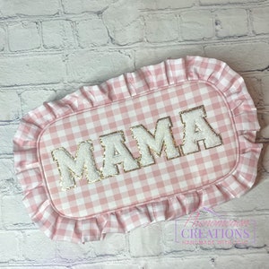May include: Pink and white gingham fabric with a ruffled edge. The fabric has the word "MAMA" embroidered in white with gold glitter accents.
