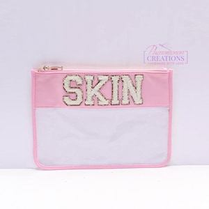 May include: A pink cosmetic bag with a clear front panel and a gold zipper. The word "SKIN" is spelled out in large, sparkly, gold-trimmed letters across the top. The bag is rectangular and has a simple, clean design.