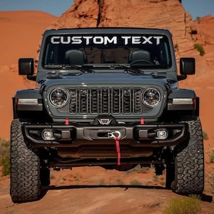 May include: A dark gray Jeep Wrangler with the text "CUSTOM TEXT" across the top of the windshield. The vehicle has large tires, a black grill, and red accents. The background features a desert landscape with red rock formations.