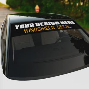 May include: A black car with a windshield decal. The decal reads "YOUR DESIGN HERE" in white and "WINDSHIELD DECAL" in gold. The car is parked outdoors.