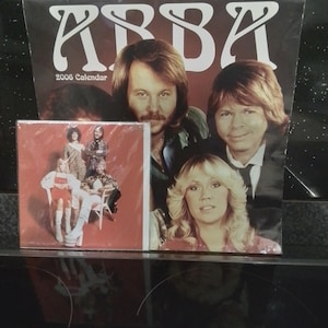 Puede incluir: Un calendario de ABBA de 2006 que presenta un gran retrato de los miembros del grupo en la portada con texto en blanco. Junto a él hay una tarjeta cuadrada más pequeña que muestra a los cuatro miembros del grupo posando sobre un fondo rojo.