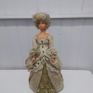 May include: A vintage doll with a blonde wig and blue eyes, dressed in a cream-coloured gown with gold detailing. The dress has lace trim and a gold glitter skirt, reminiscent of a historical era.