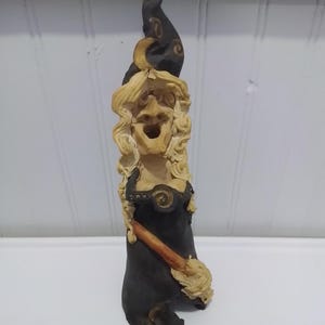 Op de afbeelding: Een lang, slank beeldje van een heks met lang, golvend blond haar, gekleed in een donkerzwarte jurk en een bijpassende puntmuts versierd met een gouden maansikkel. Het figuurtje houdt een bezem vast en heeft een wijd openstaande mond.