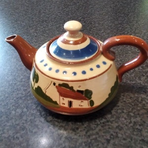 Può includere: Una teiera marrone con un coperchio a strisce bianche e blu. La teiera ha un corpo bianco con punti blu e una scena dipinta di una casa marrone con un tetto rosso e alberi verdi.