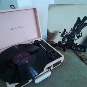 May include: A pale pink Crosley record player with a black vinyl record playing. The album cover for Kate Bush's album, "The Dreaming," is propped up beside the player. The record player is open, showing the turntable and needle.