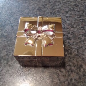 Può includere: Una scatola regalo quadrata color oro con un fiocco sulla parte superiore. Il fiocco ha un interno rosso e un piccolo abbellimento rotondo al centro. La scatola è su una superficie scura e strutturata.