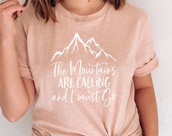 Op de afbeelding: Een t-shirt in de kleur Heather Prism Peach met een witte bergprint en de tekst "The Mountains Are Calling and I Must Go".