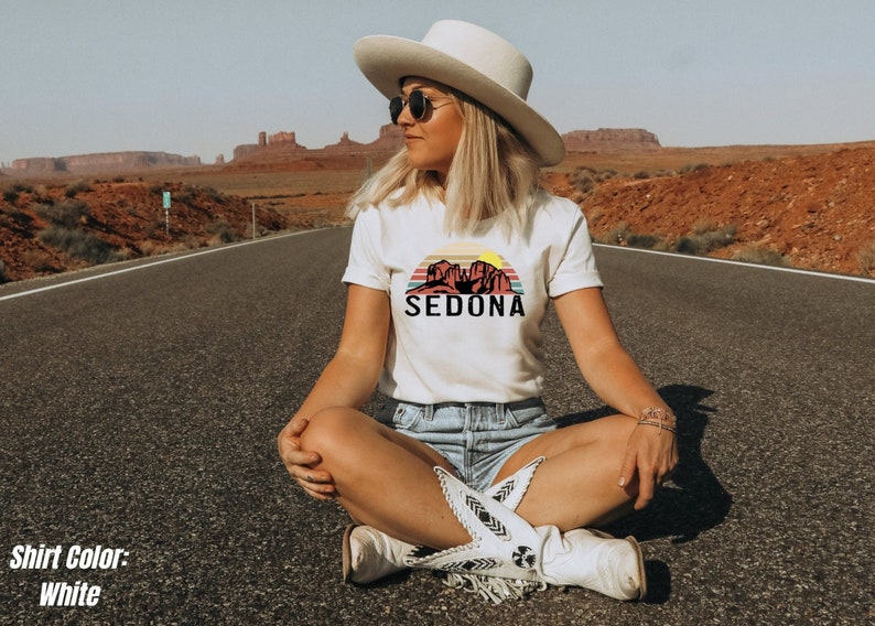 May include: A woman wearing a white t-shirt with a graphic of a sunset over red rock formations and the text "SEDONA". She is sitting on a paved road with a desert landscape in the background.