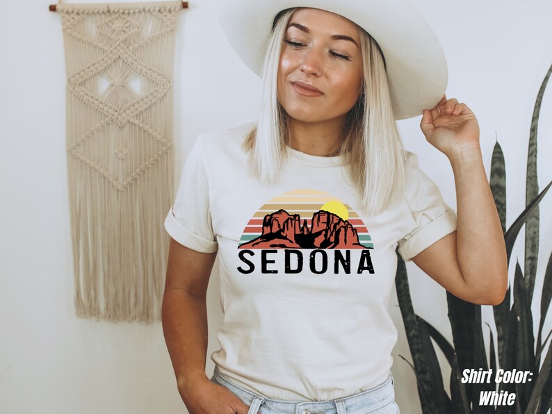 May include: A white t-shirt with a vintage-style graphic of red rock formations and a sunset with the text "SEDONA" in black.
