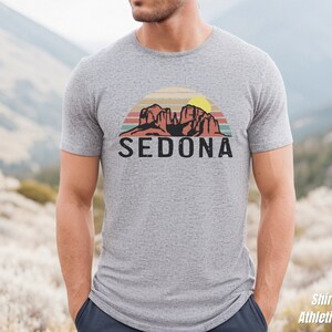 May include: A heather grey t-shirt with a retro-style graphic of red rock formations and a sunset with the word "SEDONA" printed in black below the graphic.