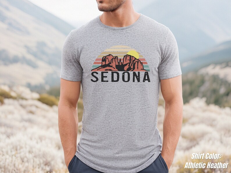 May include: A heather grey t-shirt with a retro-style graphic of red rock formations and a sunset with the word "SEDONA" printed in black below the graphic.