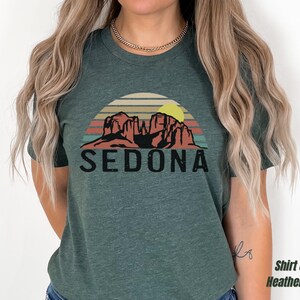 May include: A heather forest green t-shirt with a vintage-style graphic of red rock formations and a yellow sun with the text "SEDONA" in black.