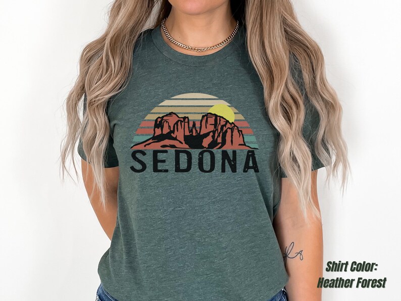 May include: A heather forest green t-shirt with a vintage-style graphic of red rock formations and a yellow sun with the text "SEDONA" in black.