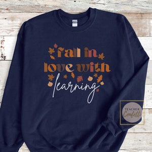 May include: Navy blue sweatshirt with the text "Fall in love with learning" in brown and orange letters with autumn leaves surrounding the text. The text "Teacher Confetti" is in a circle in the lower right corner.