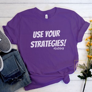 May include: Purple t-shirt with the text "USE YOUR STRATEGIES! #testday" in white.