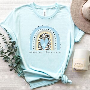 May include: A light blue t-shirt with a rainbow design featuring a heart with a puzzle piece inside. The rainbow is made of blue, yellow, and gray stripes with black dots. The text "#Autism Awareness" is printed below the rainbow.