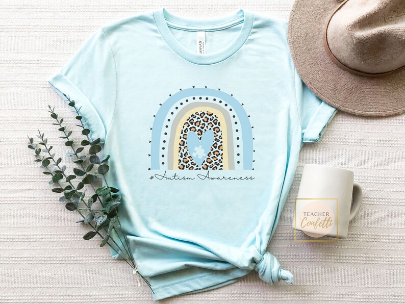 May include: A light blue t-shirt with a rainbow design featuring a heart with a puzzle piece inside. The rainbow is made of blue, yellow, and gray stripes with black dots. The text "#Autism Awareness" is printed below the rainbow.