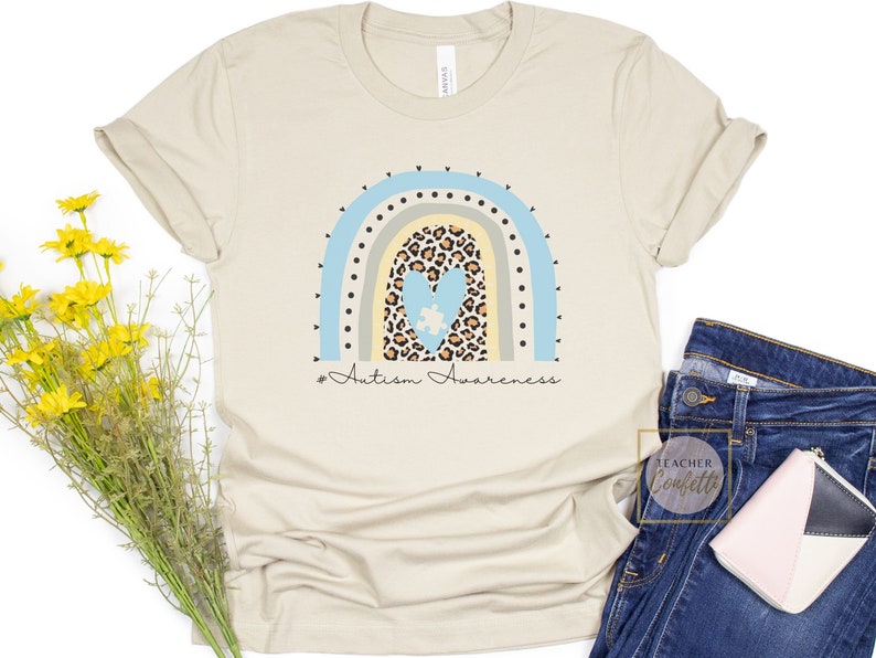 May include: A beige t-shirt with a rainbow design featuring a heart with a puzzle piece inside. The rainbow is made of blue, yellow, and green stripes with black polka dots. The text "# Autism Awareness" is printed below the rainbow.