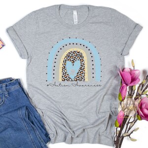 May include: A gray t-shirt with a rainbow design featuring a blue heart with a puzzle piece inside. The rainbow is made of blue, yellow, and gray stripes with black polka dots. The text "# Autism Awareness" is printed below the rainbow.