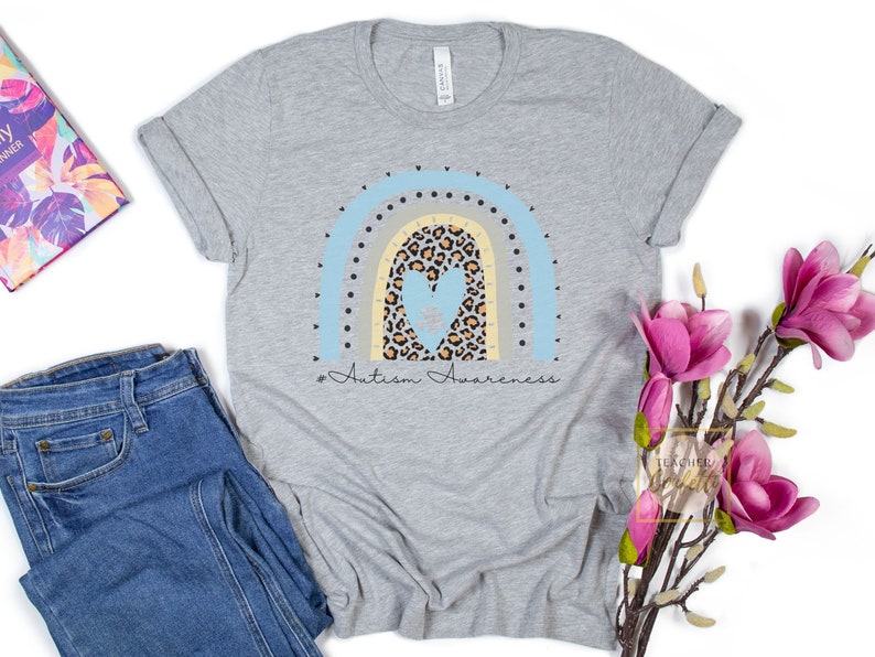May include: A gray t-shirt with a rainbow design featuring a blue heart with a puzzle piece inside. The rainbow is made of blue, yellow, and gray stripes with black polka dots. The text "# Autism Awareness" is printed below the rainbow.