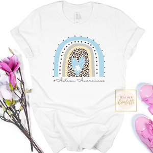 May include: White t-shirt with a blue, beige, and brown rainbow design. The rainbow has a heart with a puzzle piece inside, and the text "#Autism Awareness" is below the rainbow.