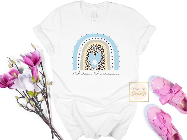 May include: White t-shirt with a blue, beige, and brown rainbow design. The rainbow has a heart with a puzzle piece inside, and the text "#Autism Awareness" is below the rainbow.