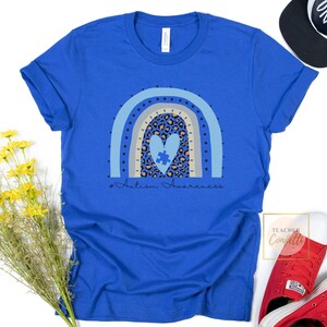 May include: A royal blue t-shirt with a rainbow design featuring a heart with a puzzle piece inside. The text "#Autism Awareness" is printed below the rainbow.