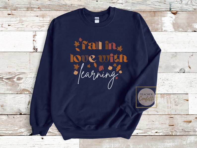 May include: Navy blue sweatshirt with the text "Fall in love with learning" in brown and orange letters with autumn leaves surrounding the text. The text "Teacher Confetti" is in a circle in the lower right corner.