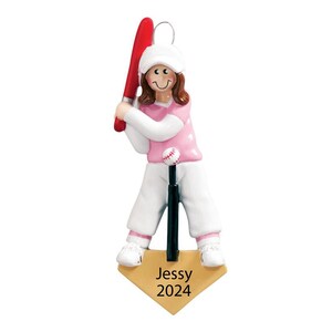 May include: A baseball-themed ornament featuring a figure in a pink shirt, white pants, and a white hat, holding a red bat. A baseball sits on a tee. The base of the ornament is a home plate shape with the name "Jessy" and the year "2024".