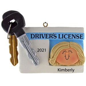 May include: A white and blue driver's license ornament with a cartoon woman's face and the text "DRIVER'S LICENSE" and "Kimberly". The ornament also includes two keys, one black and one gold.