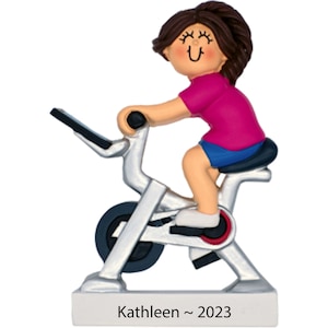 May include: A silver exercise bike with a woman figurine riding it. The figurine has brown hair, a pink shirt, blue shorts, and is smiling. The base of the bike has the text "Kathleen ~ 2023".