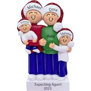 May include: A Christmas ornament depicting a family of four with red and white Santa hats. The parents are holding a baby bump and the caption reads "Expecting Again! 2023". The children are named Emma and Steven.