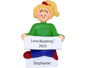 May include: A personalized Christmas ornament featuring a girl with blonde hair wearing a green sweater and blue jeans. She is holding a sign that says "Love Reading! 2023" and the name "Stephanie" is written below the sign.