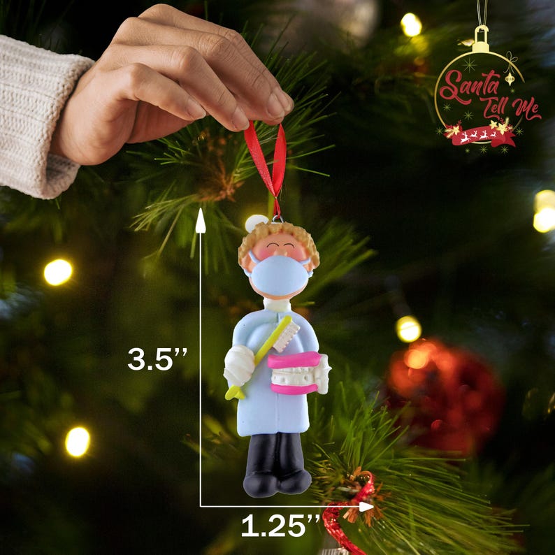 May include: A blue and white Christmas ornament depicting a dentist wearing a face mask and holding a toothbrush and a pink set of teeth. The ornament is 3.5 inches tall and 1.25 inches wide. The ornament is hanging from a red ribbon. The text "Santa Tell Me" is visible in the background.