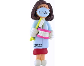 May include: A personalized Christmas ornament featuring a female dentist wearing a blue scrub top, a blue face mask with the name "Linda" written on it, and holding a toothbrush and a set of teeth. The ornament is decorated with a red skirt and the year "2022".