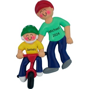 May include: A personalized Christmas ornament featuring a father and son. The father is wearing a red hat, green shirt, and blue jeans. The son is wearing a green helmet, yellow shirt, blue jeans, and riding a red tricycle. The ornament is personalized with the names "Michael 2024" and "James".