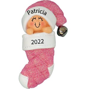 May include: Pink glitter stocking ornament with a baby girl inside wearing a Santa hat. The stocking has a white cuff with the name "Patricia" and the year "2022" written on it.