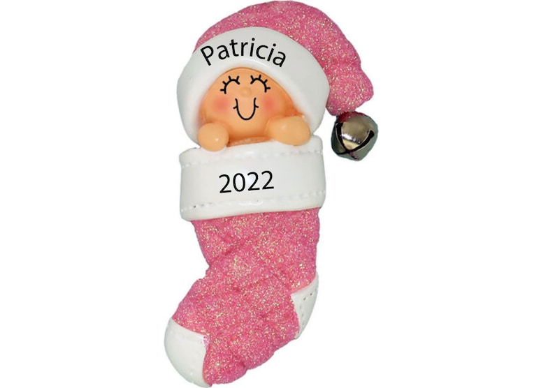 May include: Pink glitter stocking ornament with a baby girl inside wearing a Santa hat. The stocking has a white cuff with the name "Patricia" and the year "2022" written on it.