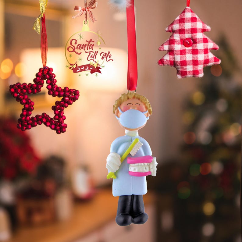 May include: A Christmas ornament featuring a cartoon dentist wearing a blue coat and a face mask, holding a toothbrush and a set of teeth. The ornament is hanging from a red ribbon. The text "Santa Tell Me" is visible in the background.