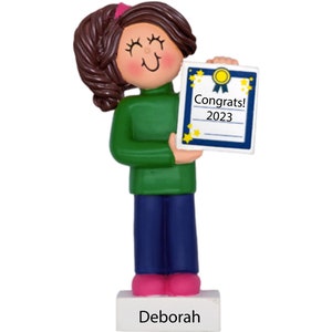 May include: A personalized ornament featuring a cartoon girl with brown hair wearing a green sweater and blue jeans. She is holding a blue and white diploma with gold stars and the text "Congrats! 2023". The ornament has a white base with the name "Deborah" written on it.