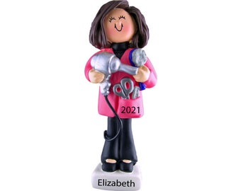 May include: A personalized Christmas ornament featuring a cartoon woman with brown hair, wearing a pink jacket and black pants. She is holding a hairdryer, scissors, and a comb. The ornament is inscribed with the name "Elizabeth" and the year "2021".