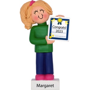 May include: A personalized ornament featuring a girl with blonde hair and a green sweater holding a blue and white certificate with the text "Congrats! 2023". The ornament has a white base with the name "Margaret" written on it.