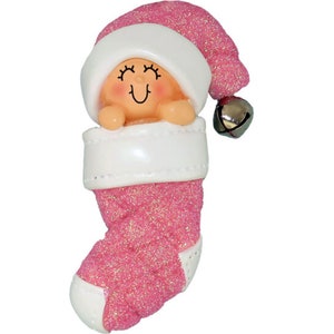 May include: Pink glitter stocking ornament with a baby doll inside wearing a white hat and a bell.