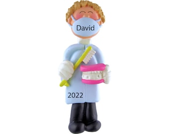 May include: A blue and white personalized ornament of a dentist holding a toothbrush and a set of dentures. The ornament has the name "David" on the face mask and the year "2022" on the bottom.