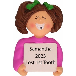 May include: A personalized Christmas ornament featuring a girl with brown hair and a missing tooth, wearing a pink shirt and holding a white sign that says "Samantha 2023 Lost 1st Tooth".