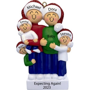 May include: A personalized Christmas ornament featuring a family of five, including a pregnant mother, a father, and three children. The ornament is decorated with red, green, and white colors and includes the names "Michael", "Dora", "Emma", "Ron", "Steven", and "Baby". The ornament also includes the text "Expecting Again! 2023".